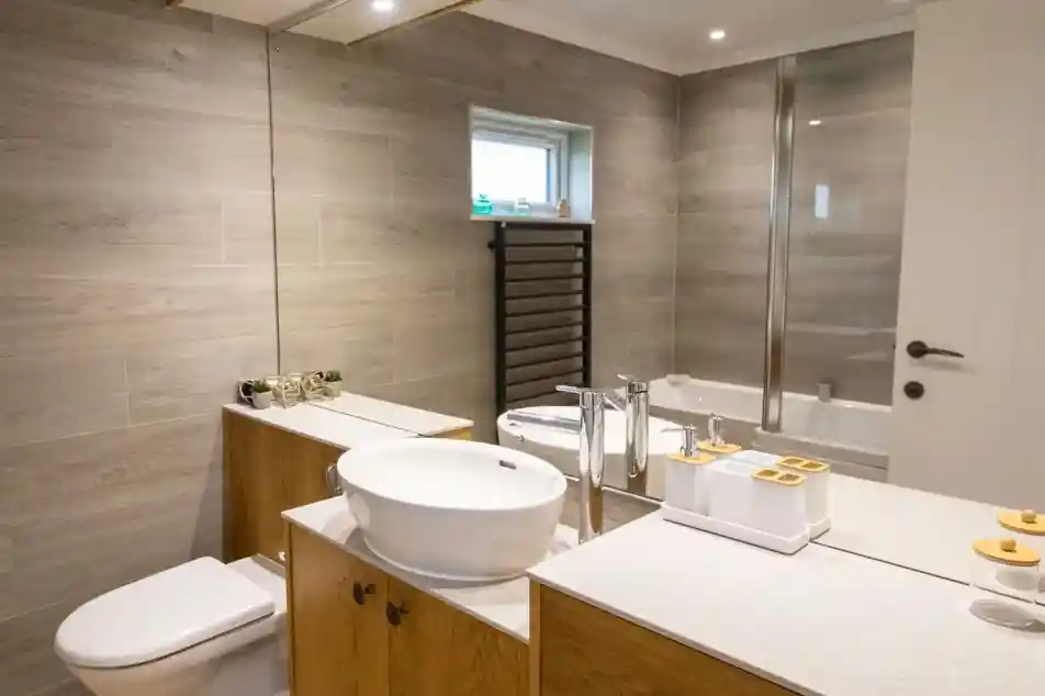 A modern bathroom features a wooden vanity with a round sink, a mirror above, and decorative items. There’s a toilet and a glass shower enclosure, along with gray tiled walls and a small window.