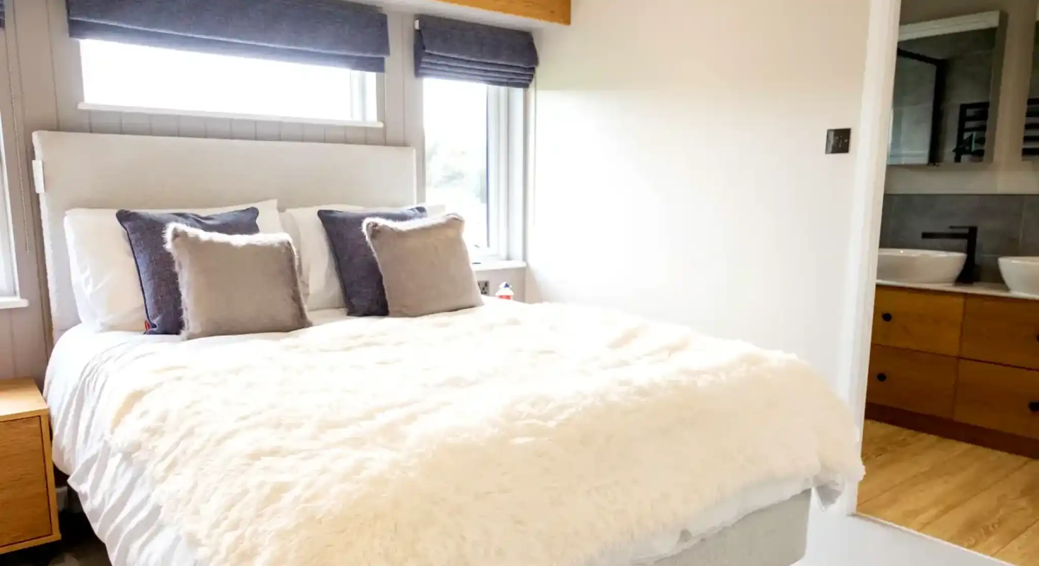 A large double bedroom featuring a plush white duvet, grey headboard, and a glimpse of a modern bathroom with double sinks.