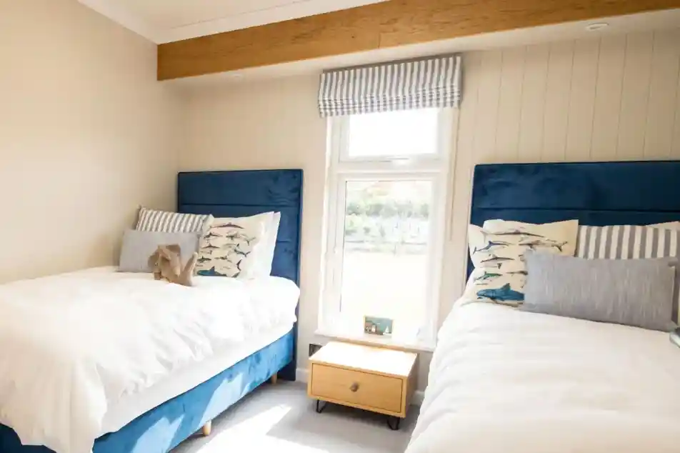 A cozy bedroom featuring two twin beds with blue upholstered headboards, dressed in white linens and decorative pillows. A small wooden nightstand sits between the beds, and a window offers a view, covered by striped curtains.