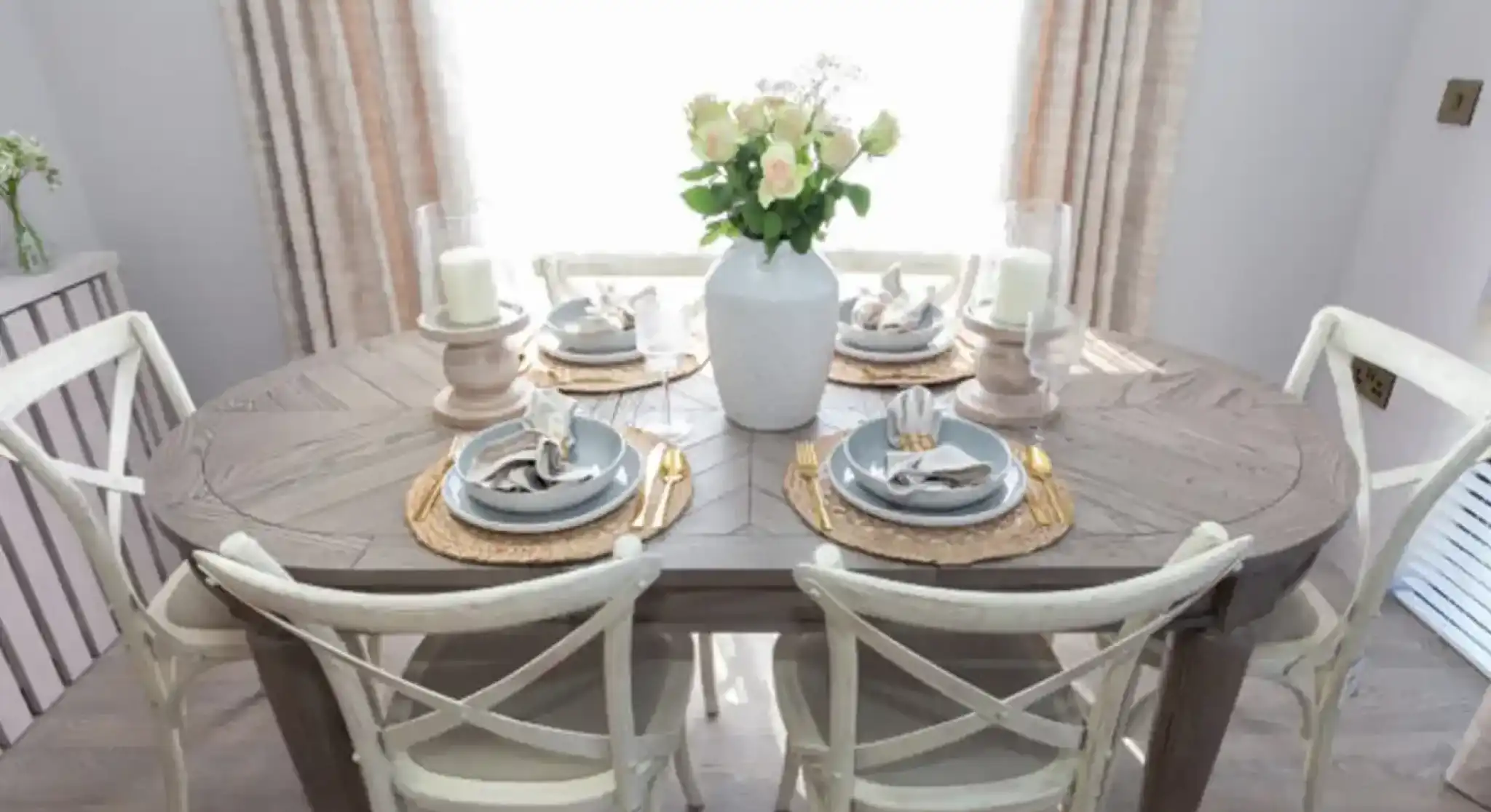 A dining table set for six with a vase of white roses and candlelight.