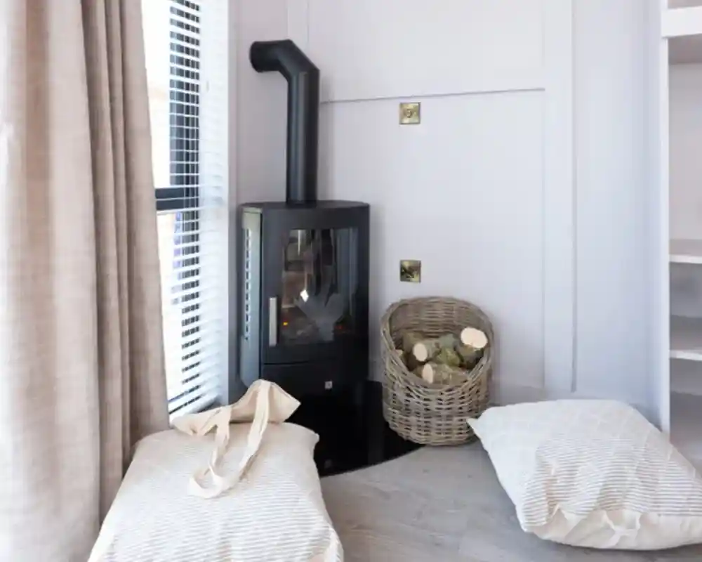 A modern wood stove is situated in a cozy corner next to a basket of firewood. Two soft, striped cushions are placed on the floor near the stove, and sunlight filters through a window with blinds.