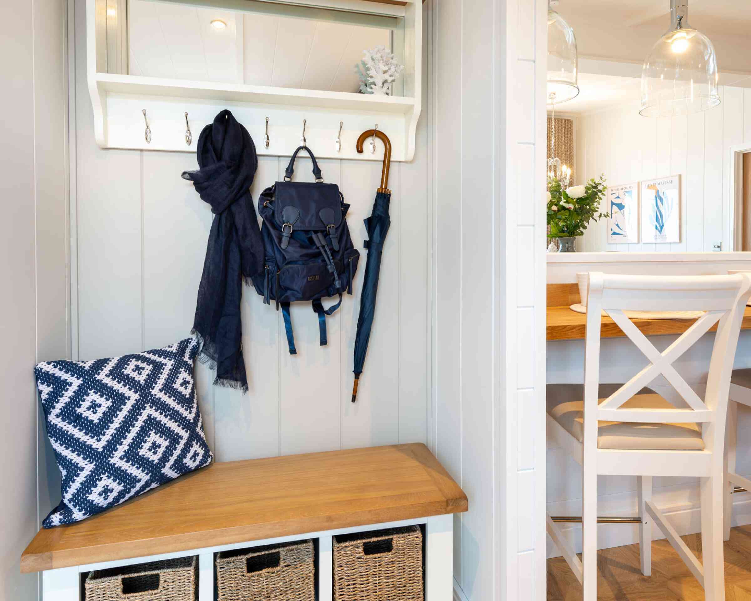 A modern entryway featuring a wooden bench with storage baskets, a navy blue backpack and scarf hanging on hooks, and a dining area with a wooden table and chairs in the background. Decorative elements include a small vase of flowers and framed artwork on the walls.