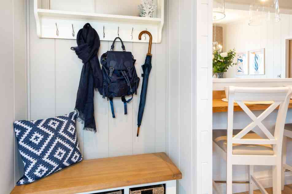 A modern entryway featuring a wooden bench with storage baskets, a navy blue backpack and scarf hanging on hooks, and a dining area with a wooden table and chairs in the background. Decorative elements include a small vase of flowers and framed artwork on the walls.