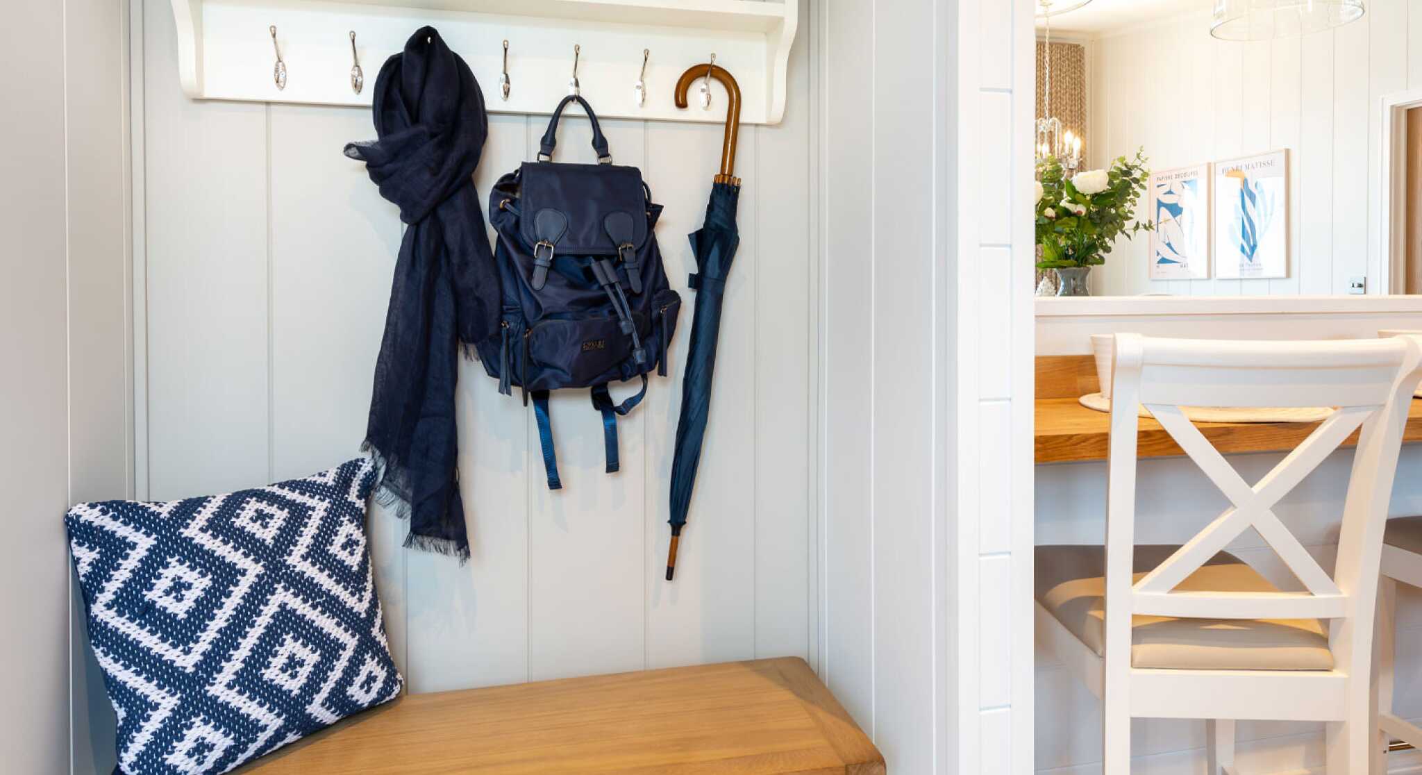 A modern entryway featuring a wooden bench with storage baskets, a navy blue backpack and scarf hanging on hooks, and a dining area with a wooden table and chairs in the background. Decorative elements include a small vase of flowers and framed artwork on the walls.