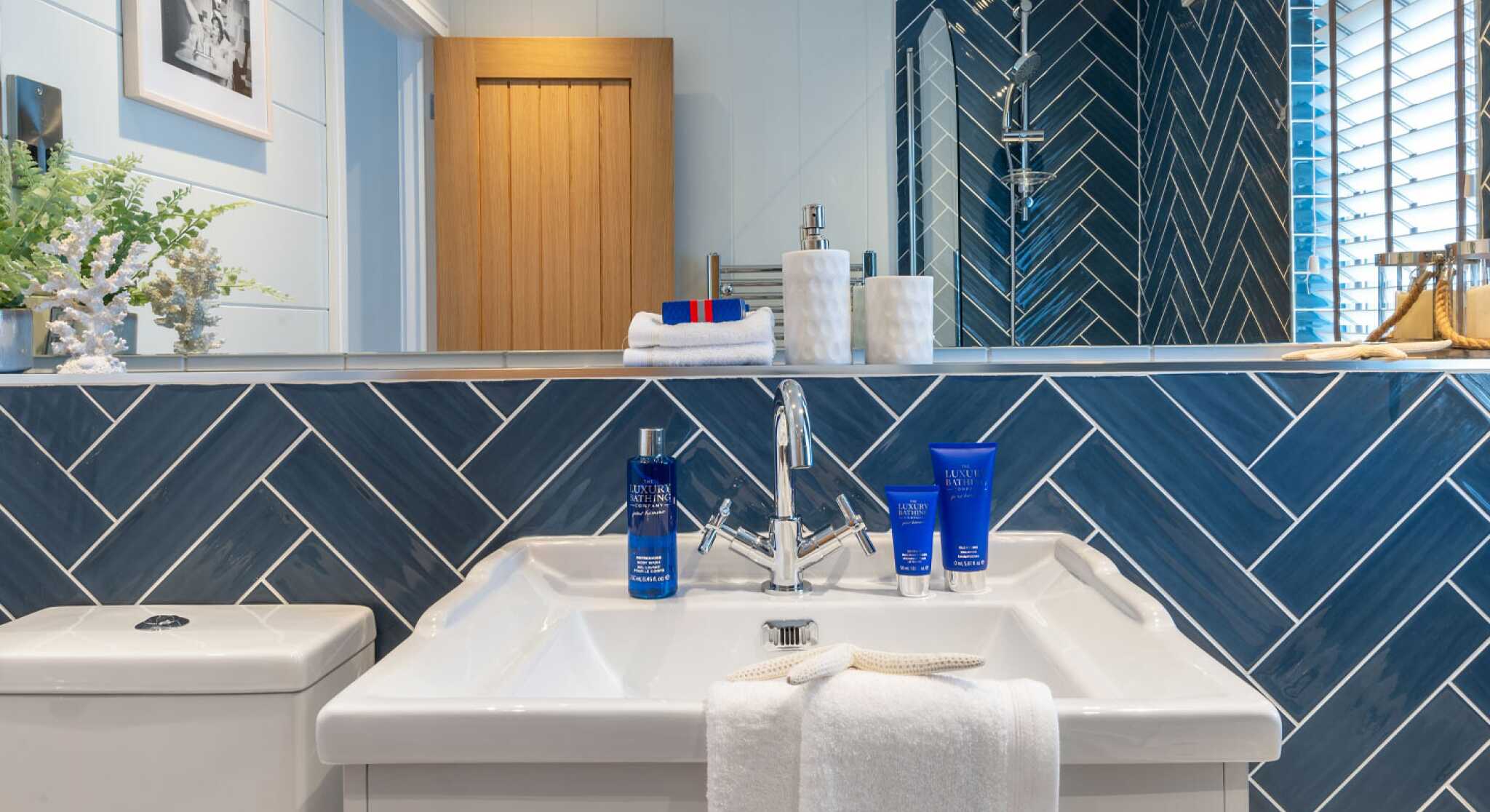 Modern bathroom with a herringbone-patterned blue tiled wall. A white sink with a sleek faucet is displayed, topped with a towel and blue skincare products. Natural light illuminates the space, highlighting the wooden door and decorative elements.