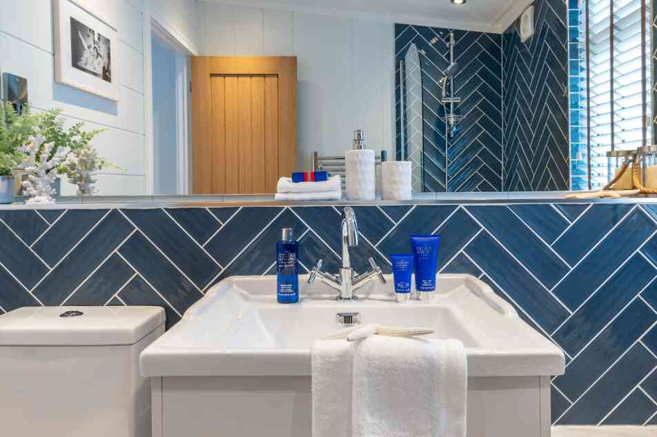 Modern bathroom with a herringbone-patterned blue tiled wall. A white sink with a sleek faucet is displayed, topped with a towel and blue skincare products. Natural light illuminates the space, highlighting the wooden door and decorative elements.