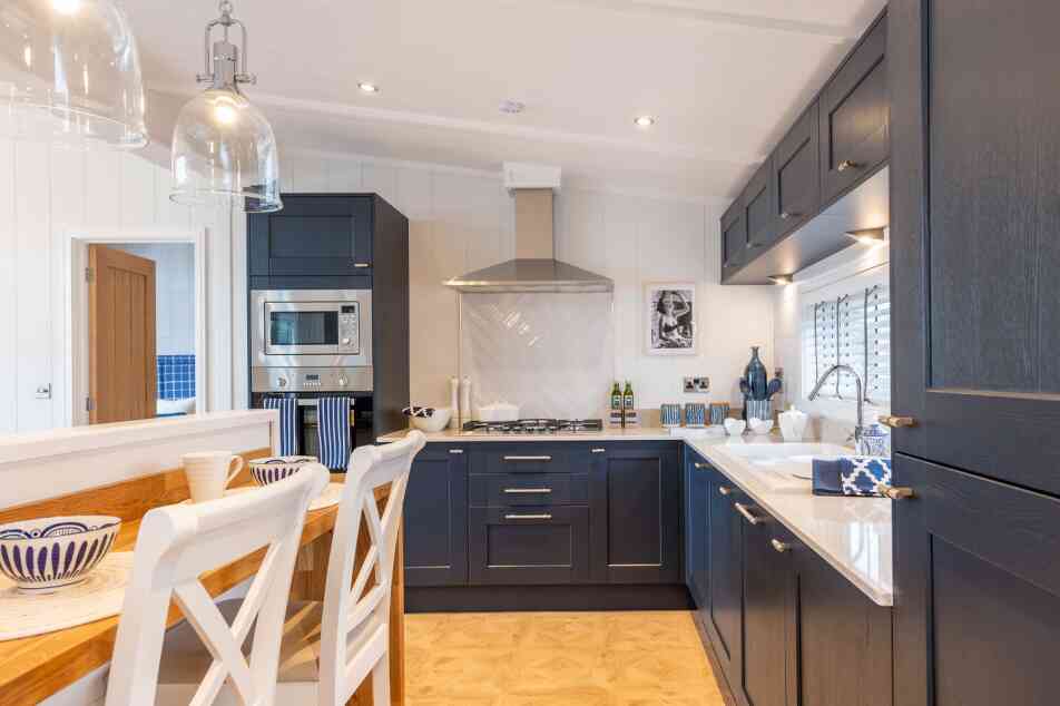 Modern kitchen featuring dark blue cabinetry, a stainless steel range hood, and a spacious countertop. A wooden dining table with white chairs is visible, and decorative items are neatly arranged on the counter. The space is well-lit with pendant lights above the table and windows allowing natural light.