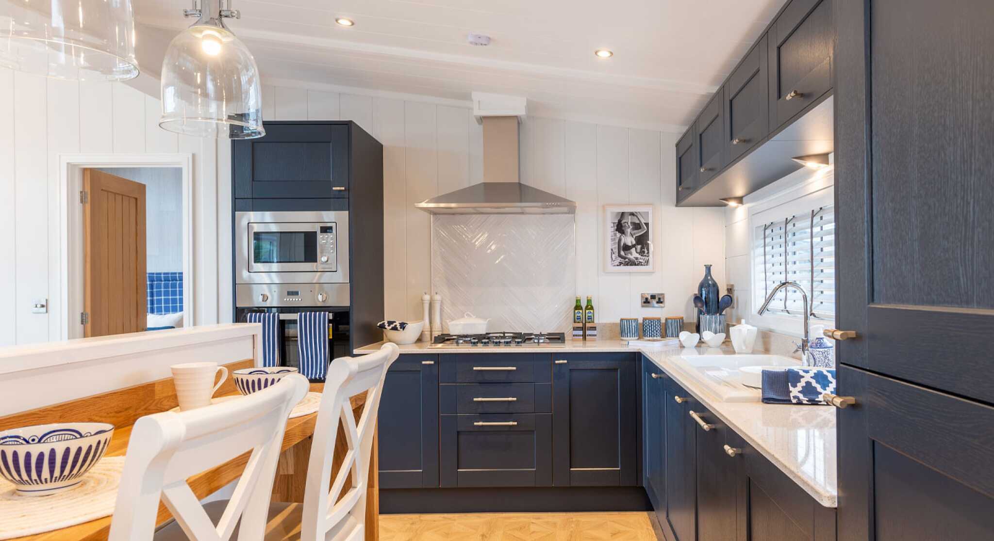 Modern kitchen featuring dark blue cabinetry, a stainless steel range hood, and a spacious countertop. A wooden dining table with white chairs is visible, and decorative items are neatly arranged on the counter. The space is well-lit with pendant lights above the table and windows allowing natural light.