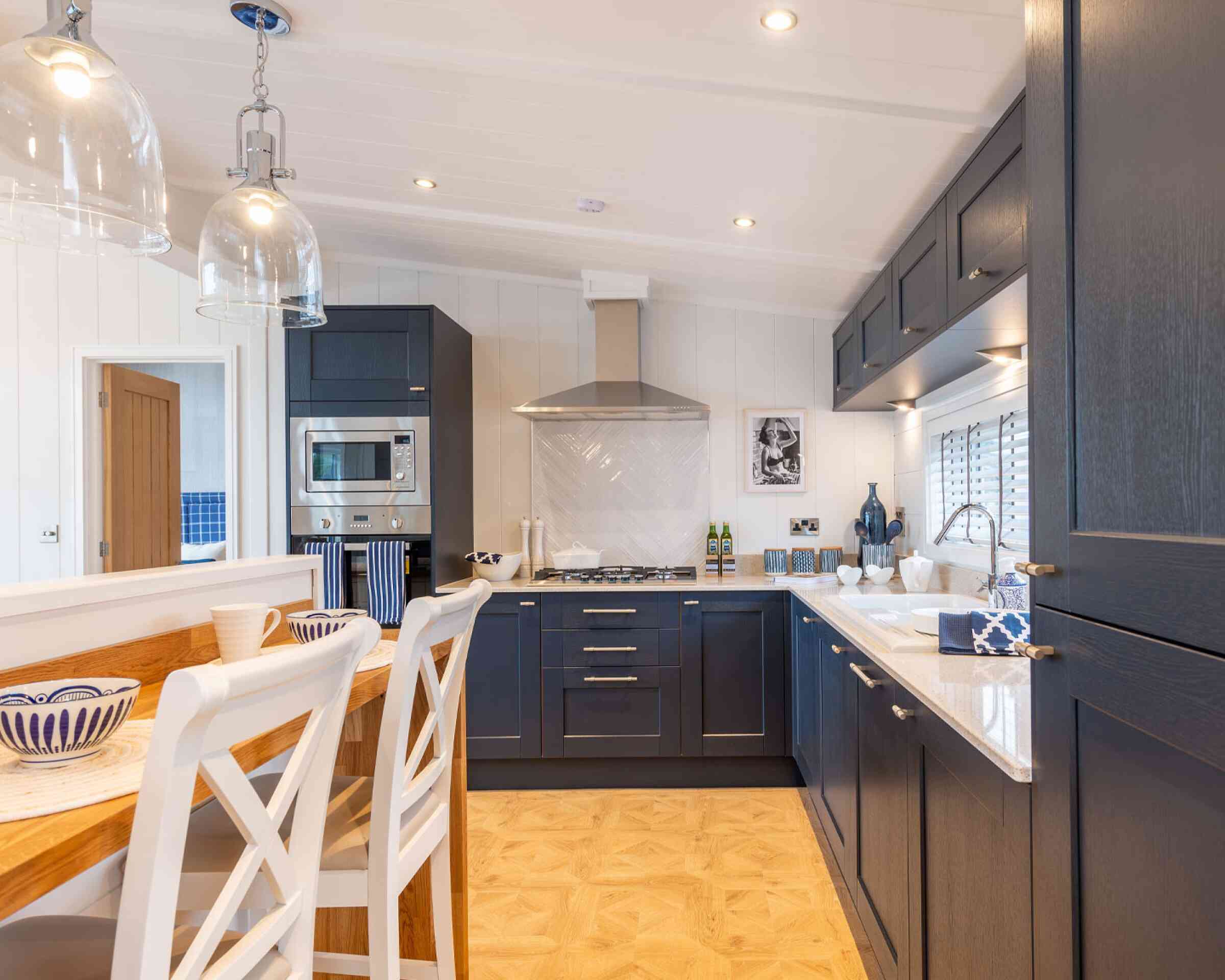 Modern kitchen featuring dark blue cabinetry, a stainless steel range hood, and a spacious countertop. A wooden dining table with white chairs is visible, and decorative items are neatly arranged on the counter. The space is well-lit with pendant lights above the table and windows allowing natural light.