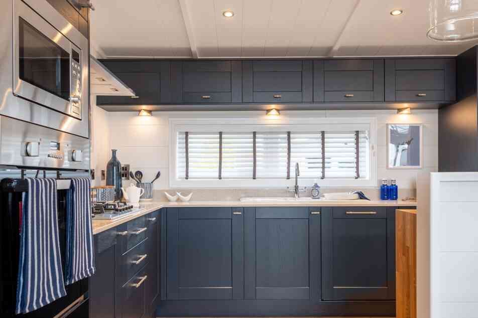 Modern kitchen featuring dark navy blue cabinetry, silver hardware, and a light countertop. Equipped with built-in appliances, including a microwave and oven. Natural light streams in through a window with white shutters, illuminating the space.