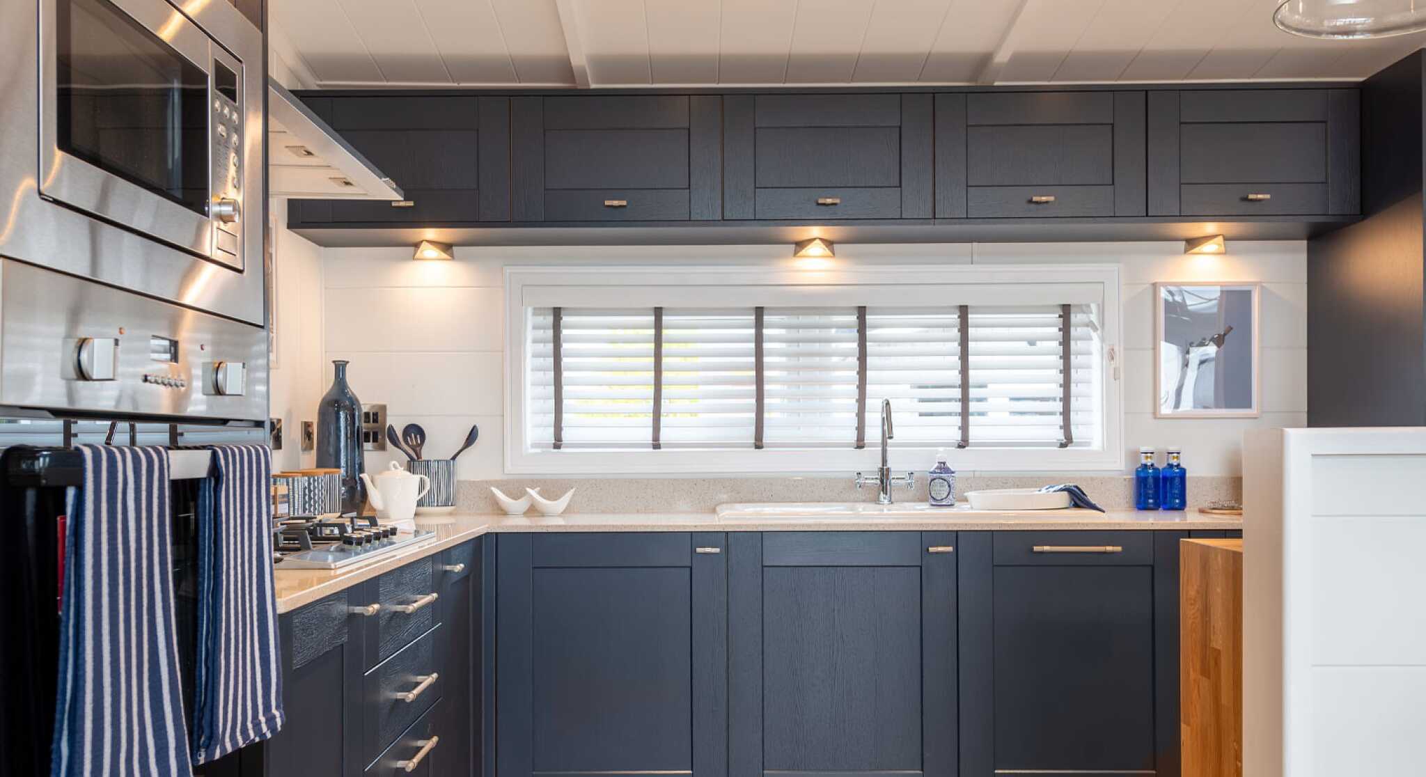 Modern kitchen featuring dark navy blue cabinetry, silver hardware, and a light countertop. Equipped with built-in appliances, including a microwave and oven. Natural light streams in through a window with white shutters, illuminating the space.