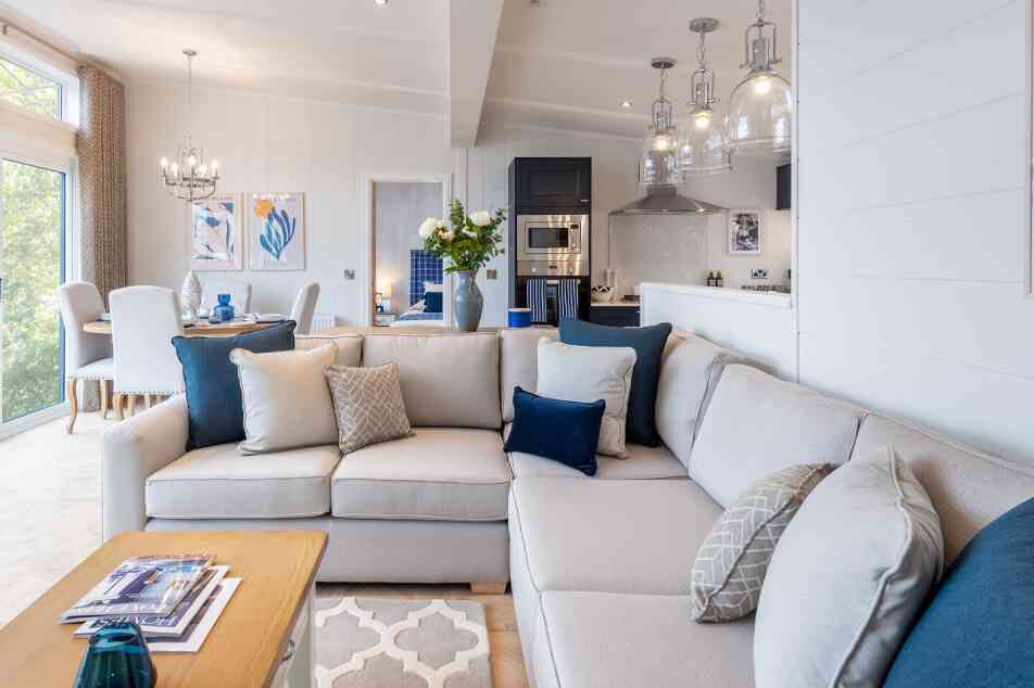 A modern living room with a large light-colored sectional sofa adorned with decorative pillows in various shades of blue and gray. A wooden coffee table sits in front, featuring magazines and a blue decorative item. The space includes stylish light fixtures, a dining area in the background, and large windows allowing natural light to fill the room.
