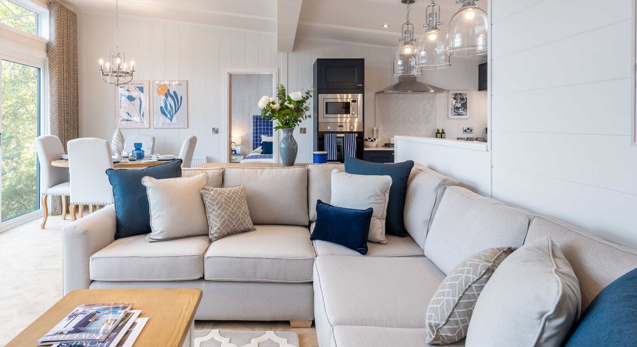 A modern living room with a large light-colored sectional sofa adorned with decorative pillows in various shades of blue and gray. A wooden coffee table sits in front, featuring magazines and a blue decorative item. The space includes stylish light fixtures, a dining area in the background, and large windows allowing natural light to fill the room.