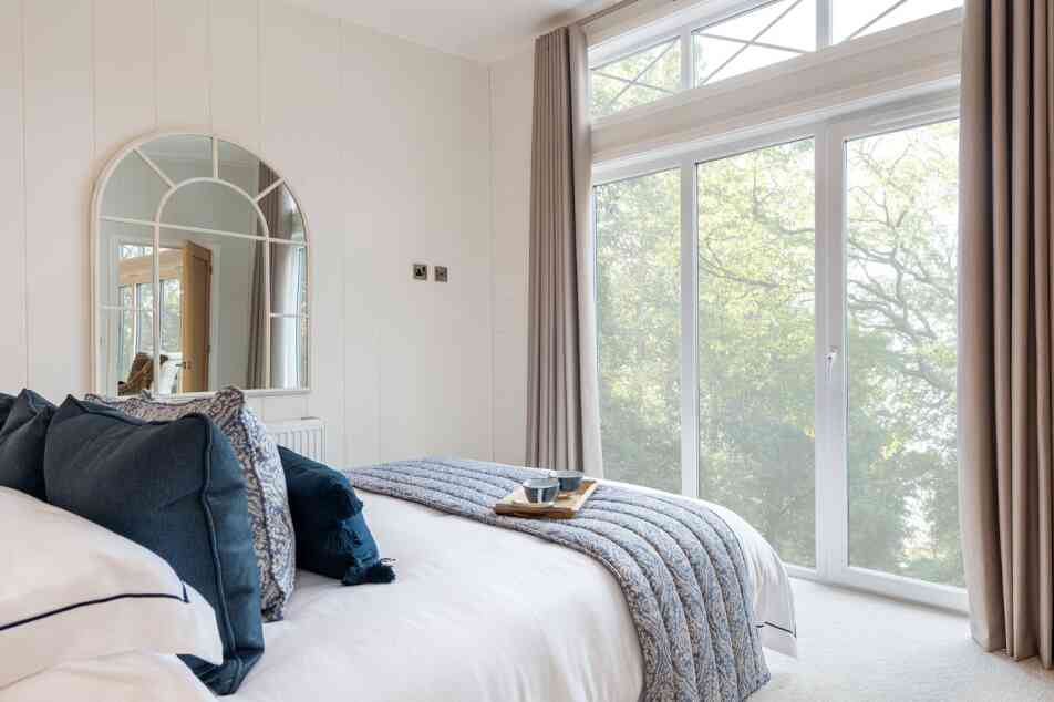 A cozy bedroom featuring a neatly made bed with navy and white bedding, a decorative tray on the bed with a cup, and large windows allowing natural light in. The walls are light-colored, and a round mirror hangs above the headboard. Soft curtains frame the windows, offering a view of greenery outside.