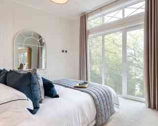 A cozy bedroom featuring a neatly made bed with navy and white bedding, a decorative tray on the bed with a cup, and large windows allowing natural light in. The walls are light-colored, and a round mirror hangs above the headboard. Soft curtains frame the windows, offering a view of greenery outside.