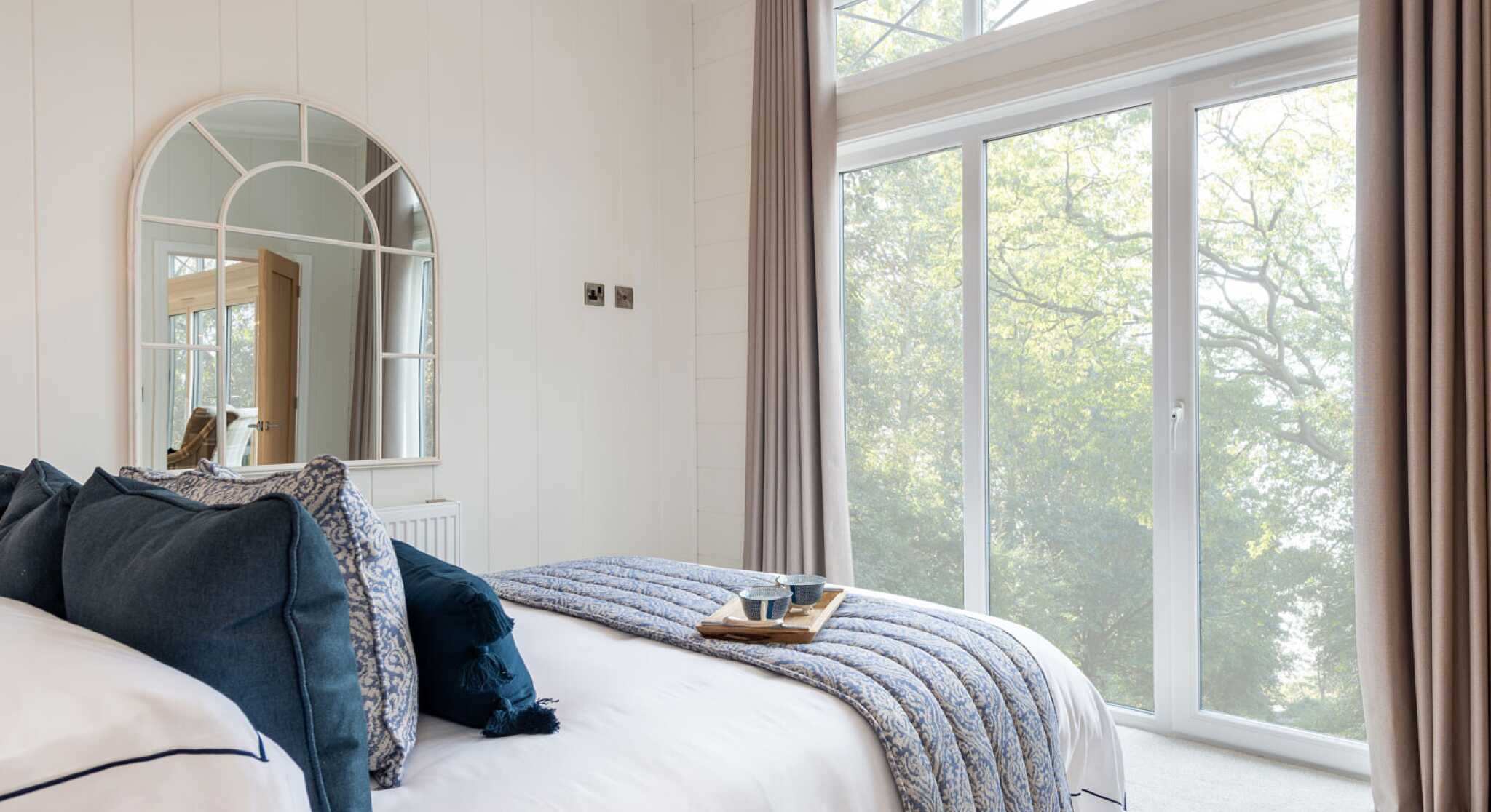 A cozy bedroom featuring a neatly made bed with navy and white bedding, a decorative tray on the bed with a cup, and large windows allowing natural light in. The walls are light-colored, and a round mirror hangs above the headboard. Soft curtains frame the windows, offering a view of greenery outside.