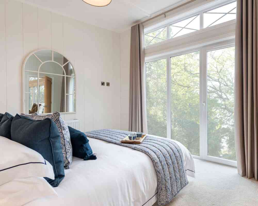 A cozy bedroom featuring a neatly made bed with navy and white bedding, a decorative tray on the bed with a cup, and large windows allowing natural light in. The walls are light-colored, and a round mirror hangs above the headboard. Soft curtains frame the windows, offering a view of greenery outside.