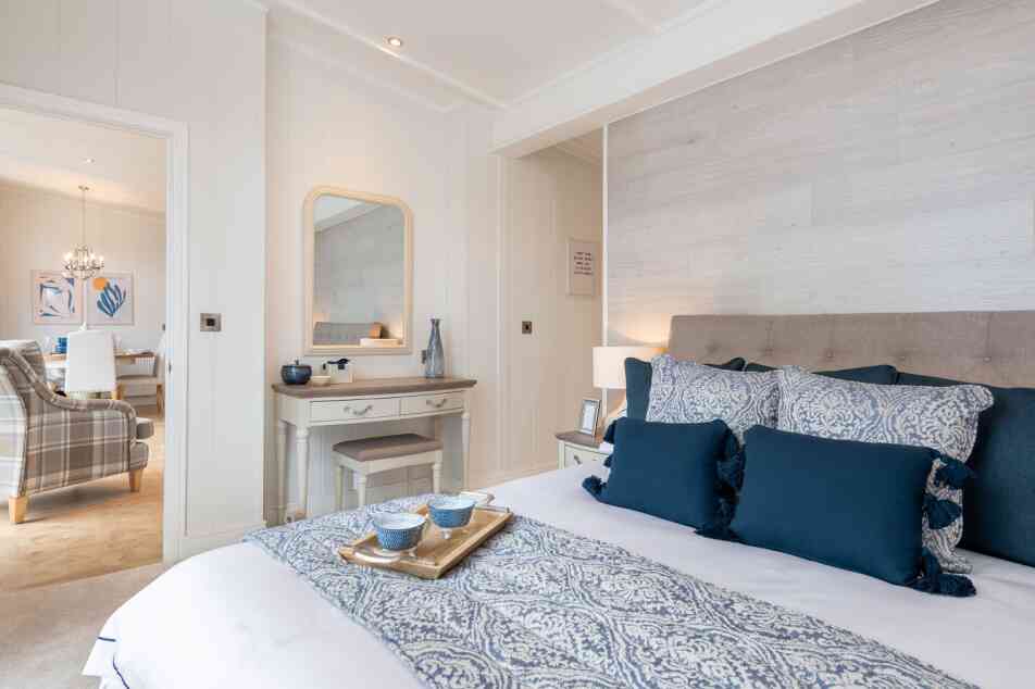 A cozy bedroom featuring a neatly made bed with blue and white patterned bedding and decorative pillows. A wooden dresser with a mirror is nearby, and a living area can be seen in the background with a chandelier and plaid sofa. Soft lighting enhances the inviting atmosphere.