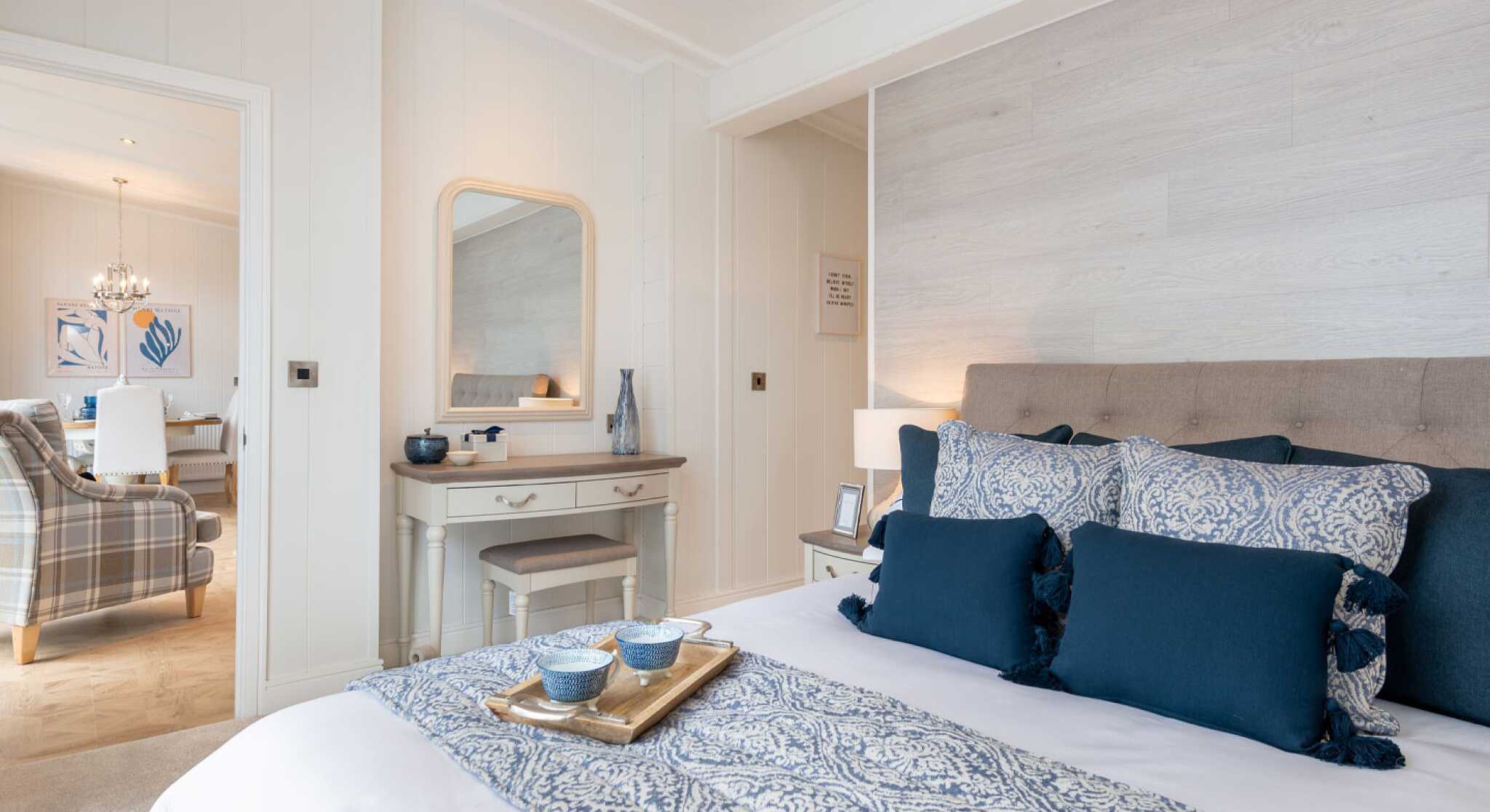 A cozy bedroom featuring a neatly made bed with blue and white patterned bedding and decorative pillows. A wooden dresser with a mirror is nearby, and a living area can be seen in the background with a chandelier and plaid sofa. Soft lighting enhances the inviting atmosphere.