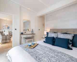 A cozy bedroom featuring a neatly made bed with blue and white patterned bedding and decorative pillows. A wooden dresser with a mirror is nearby, and a living area can be seen in the background with a chandelier and plaid sofa. Soft lighting enhances the inviting atmosphere.