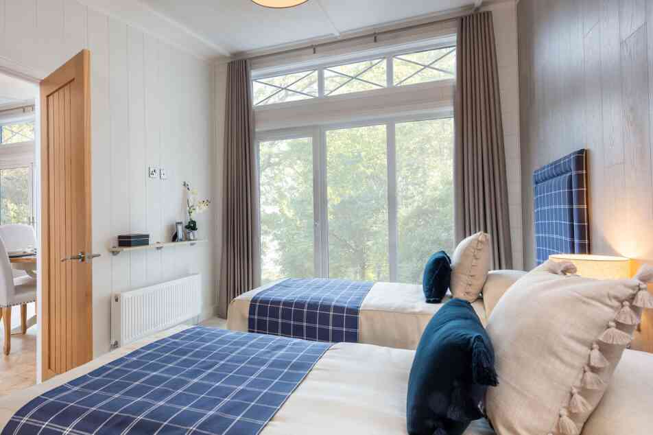 A cozy bedroom featuring two twin beds with navy blue checkered headboards. Soft, neutral bedding is complemented by decorative pillows. Large windows with sheer curtains allow natural light, with a view of greenery outside. Modern decor includes a lamp and minimalistic furniture.