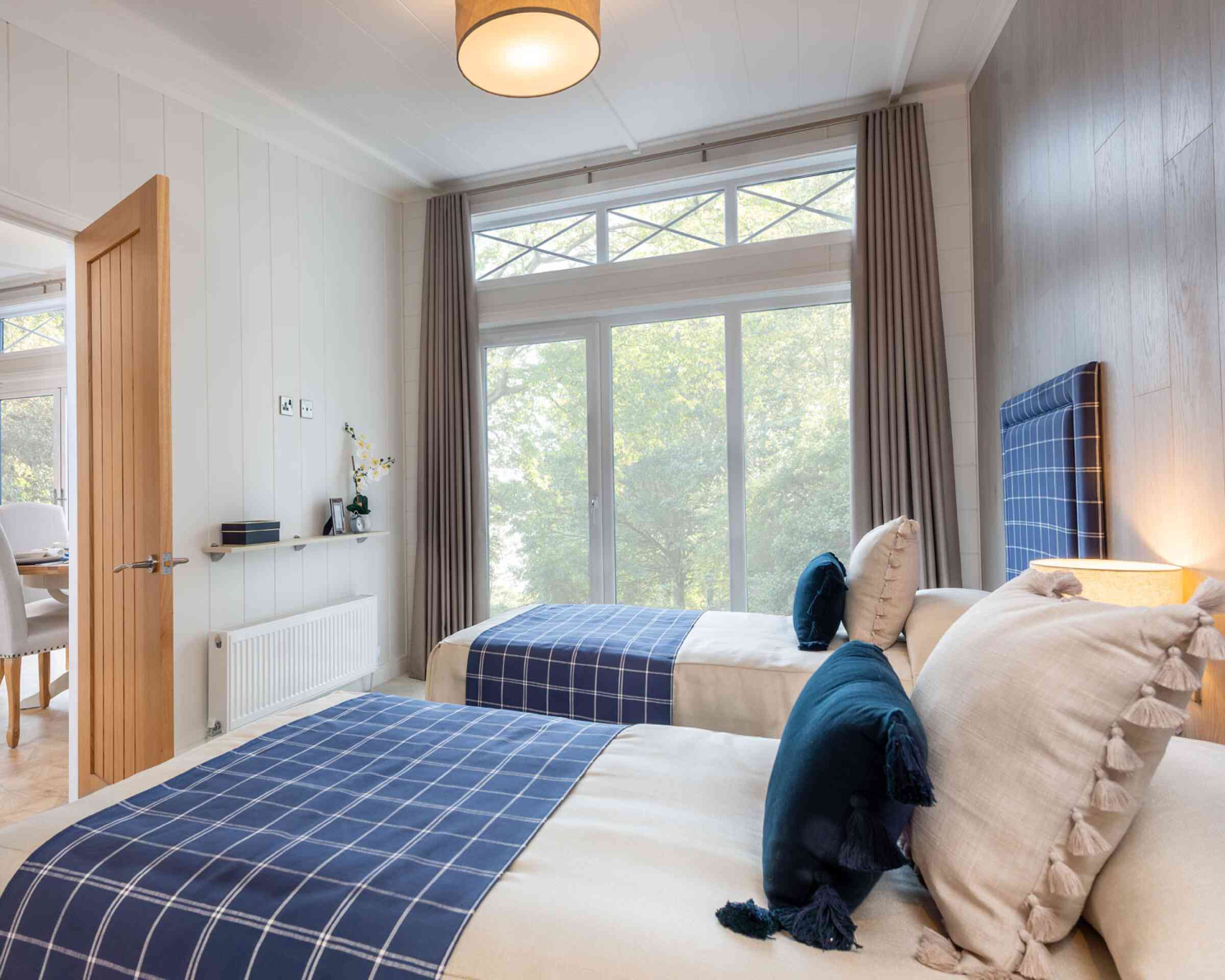 A cozy bedroom featuring two twin beds with navy blue checkered headboards. Soft, neutral bedding is complemented by decorative pillows. Large windows with sheer curtains allow natural light, with a view of greenery outside. Modern decor includes a lamp and minimalistic furniture.