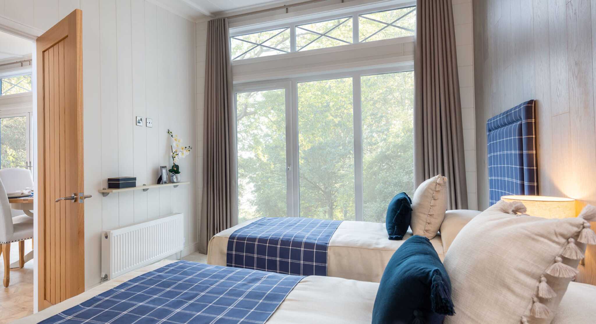 A cozy bedroom featuring two twin beds with navy blue checkered headboards. Soft, neutral bedding is complemented by decorative pillows. Large windows with sheer curtains allow natural light, with a view of greenery outside. Modern decor includes a lamp and minimalistic furniture.