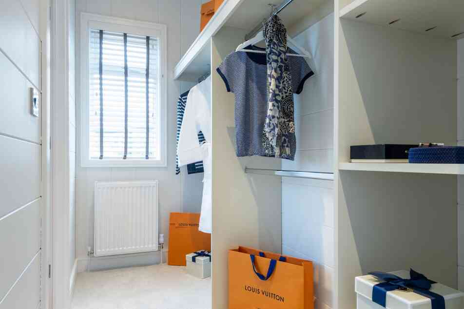 A well-organized walk-in closet featuring hanging clothes, including striped and floral patterns. The space includes shelves with neatly arranged boxes and shopping bags, notably one from Louis Vuitton. A window provides natural light, illuminating the soft carpeted floor.