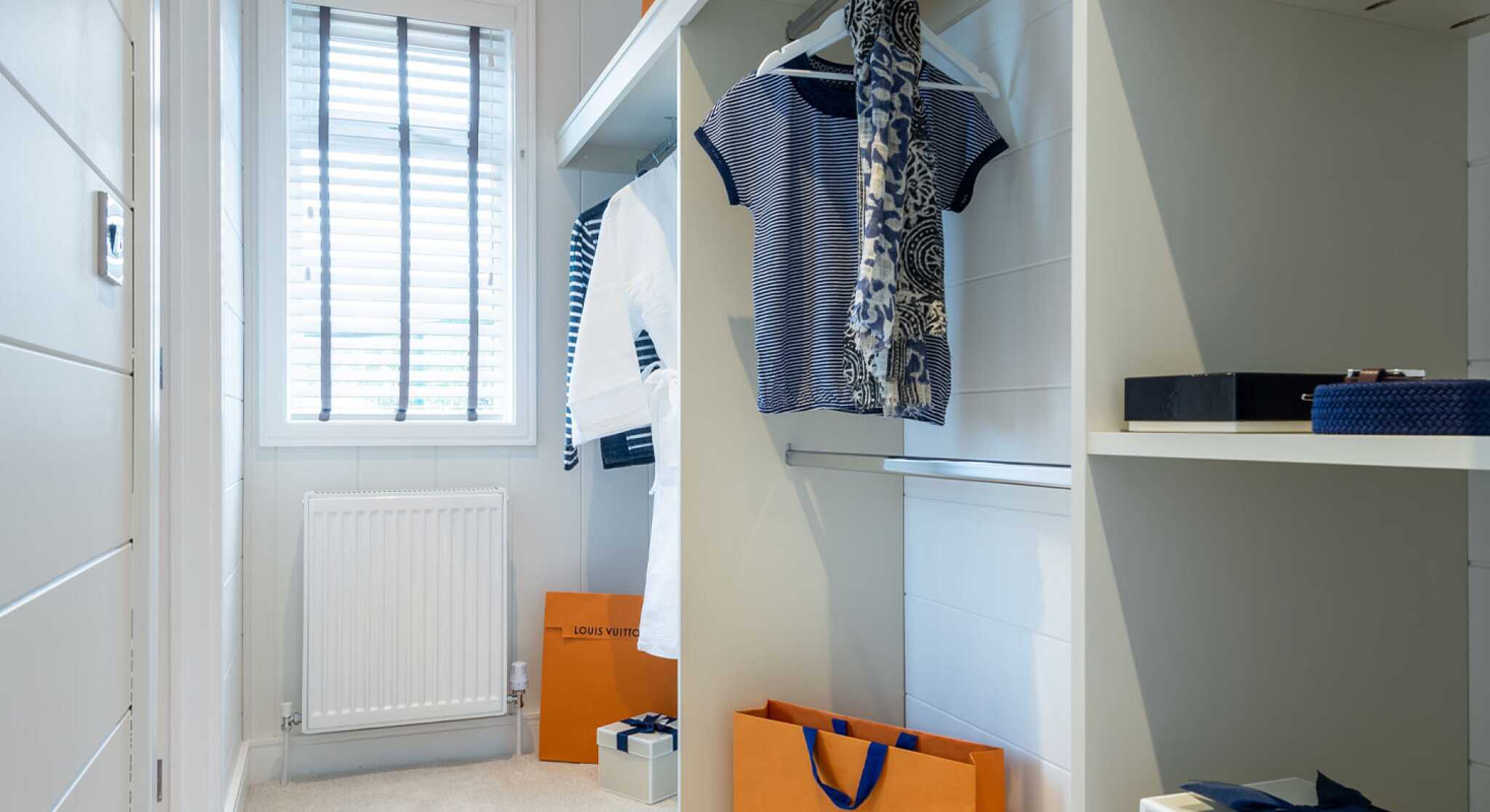 A well-organized walk-in closet featuring hanging clothes, including striped and floral patterns. The space includes shelves with neatly arranged boxes and shopping bags, notably one from Louis Vuitton. A window provides natural light, illuminating the soft carpeted floor.