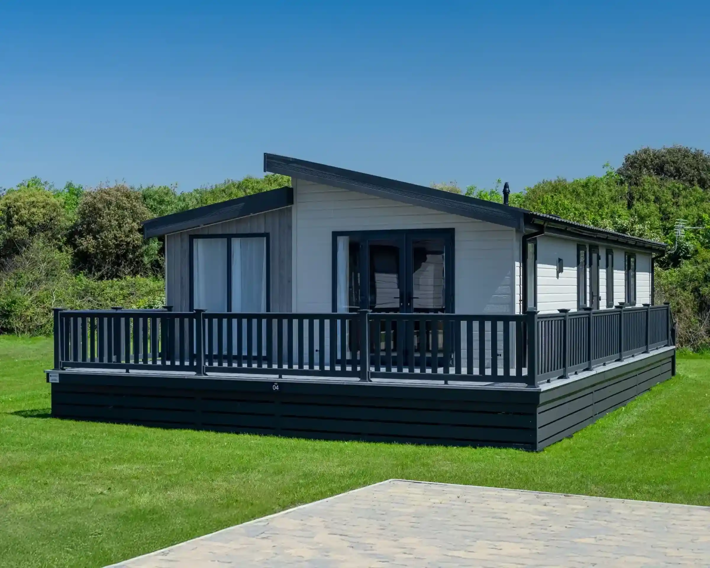 Modern white lodge holiday home with a dark wrap-around deck and grey pitched roof set on a manicured lawn.