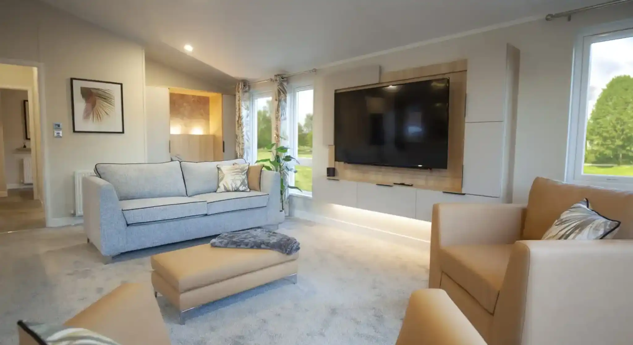 Modern living room with a large wall-mounted TV, a beige armchair, a matching ottoman, a light grey sofa with decorative pillows, and floor-to-ceiling windows allowing natural light inside. The room has neutral-toned walls, carpet flooring, recessed lighting, and a framed wall art piece.