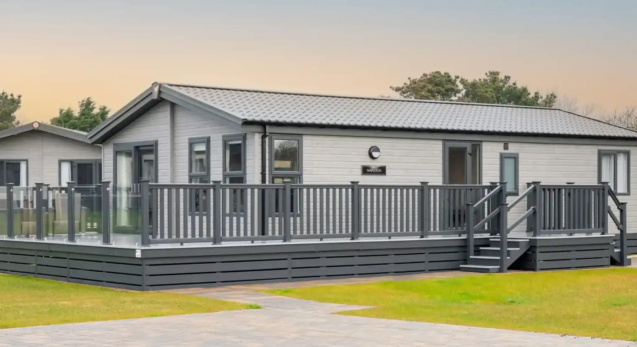 A modern, light grey lodge holiday home with a wrap-around deck and glass railings sits on a grassy lawn with a paved walkway.