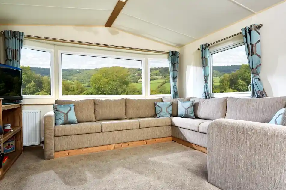 The image shows a cozy and well-lit living area, featuring a sectional sofa with soft, neutral tones and geometric accent pillows. The room has large windows offering a scenic view of a lush landscape with rolling hills, trees, and a clear sky. The space feels open and airy, with modern curtains that complement the sofa, creating a calm and inviting atmosphere. There’s also a TV unit in the corner, adding to the comfortable, relaxed vibe of the room. The soft carpet and wood elements enhance the homely, rustic charm.
