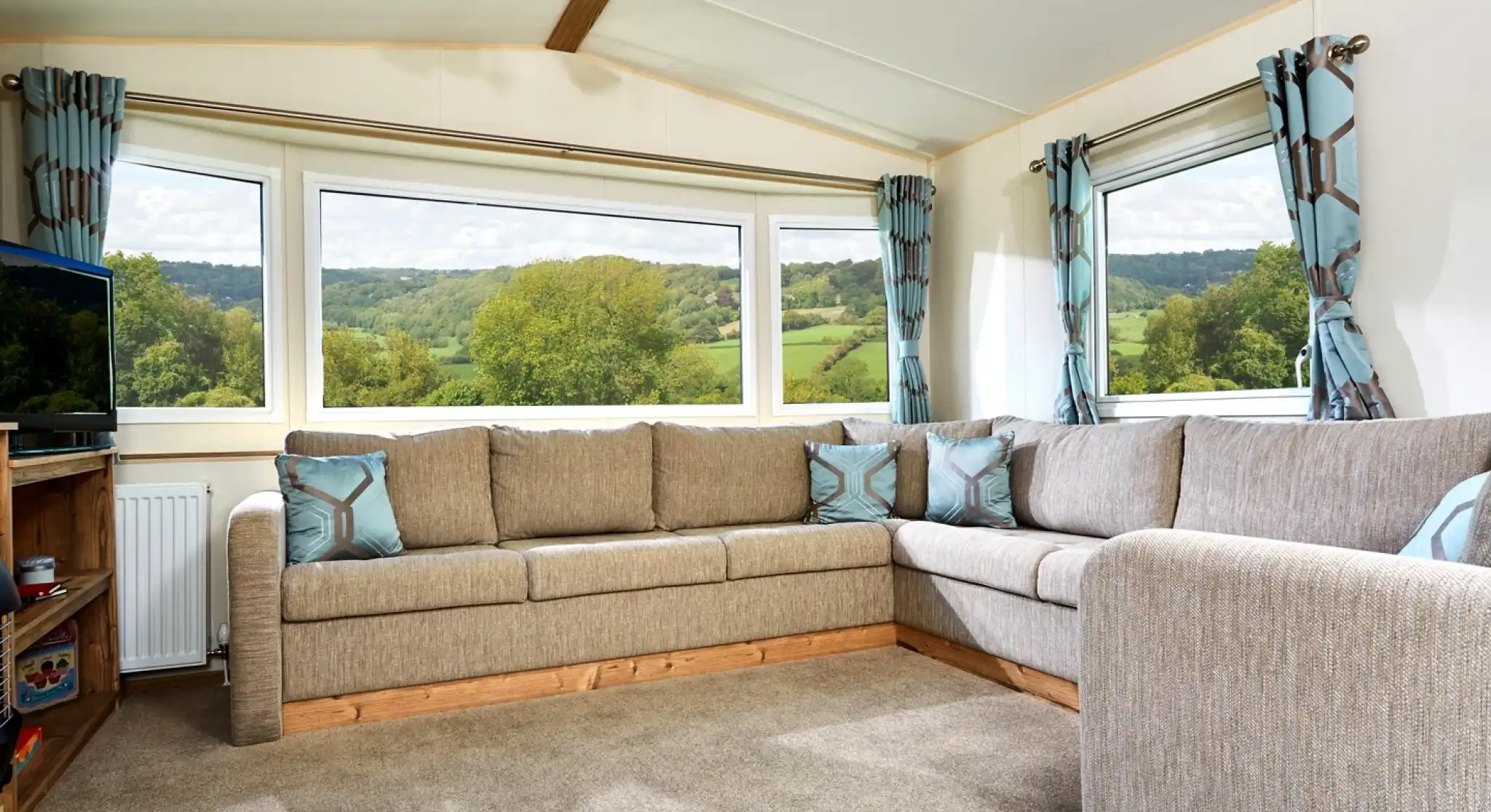 The image shows a cozy and well-lit living area, featuring a sectional sofa with soft, neutral tones and geometric accent pillows. The room has large windows offering a scenic view of a lush landscape with rolling hills, trees, and a clear sky. The space feels open and airy, with modern curtains that complement the sofa, creating a calm and inviting atmosphere. There’s also a TV unit in the corner, adding to the comfortable, relaxed vibe of the room. The soft carpet and wood elements enhance the homely, rustic charm.