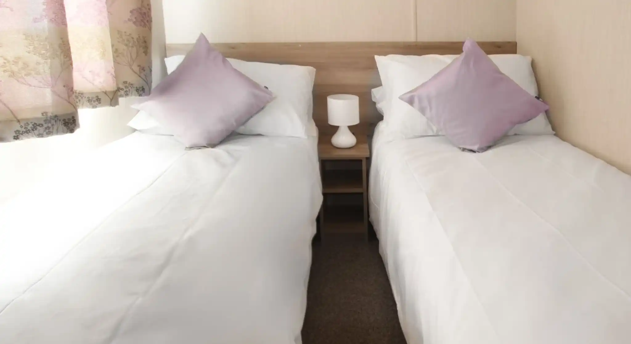 This image depicts another twin bedroom, similarly designed with a clean and simple aesthetic. The beds are neatly made with white linens and complemented by soft lavender-colored throw pillows. A small bedside table with a lamp sits between the beds, adding a cozy and functional touch. The room is bathed in natural light coming through a window with soft, patterned curtains. The neutral tones throughout the room contribute to a tranquil and inviting atmosphere.