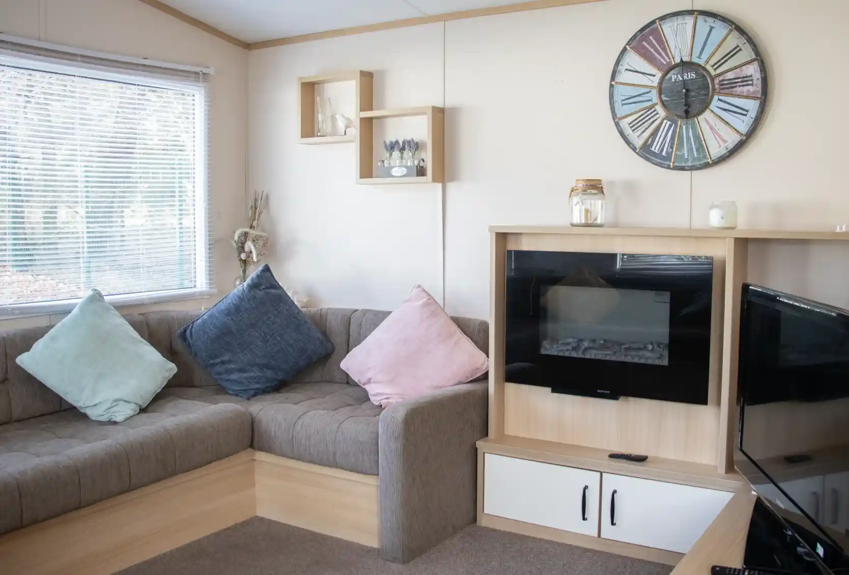 A cozy living room with a brown sectional sofa adorned with blue and pink cushions. A modern clock hangs on the wall, and there's a small shelf with decorative items. A fireplace is built into a wooden cabinet, next to a TV stand. Natural light filters through a window with blinds.