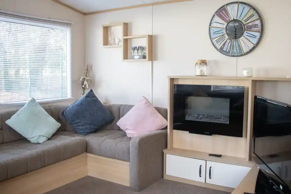 A cozy living room with a brown sectional sofa adorned with blue and pink cushions. A modern clock hangs on the wall, and there's a small shelf with decorative items. A fireplace is built into a wooden cabinet, next to a TV stand. Natural light filters through a window with blinds.