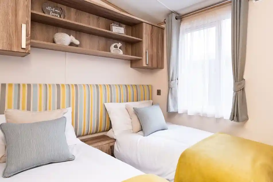 Bright and cozy twin bedroom with two single beds featuring white bedding, yellow throws, and striped yellow and gray headboards. Each bed has a gray and beige cushion. A window with sheer white curtains and gray drapes lets in natural light. Above the beds, there are wooden cabinets and open shelving with decorative items and ornaments.