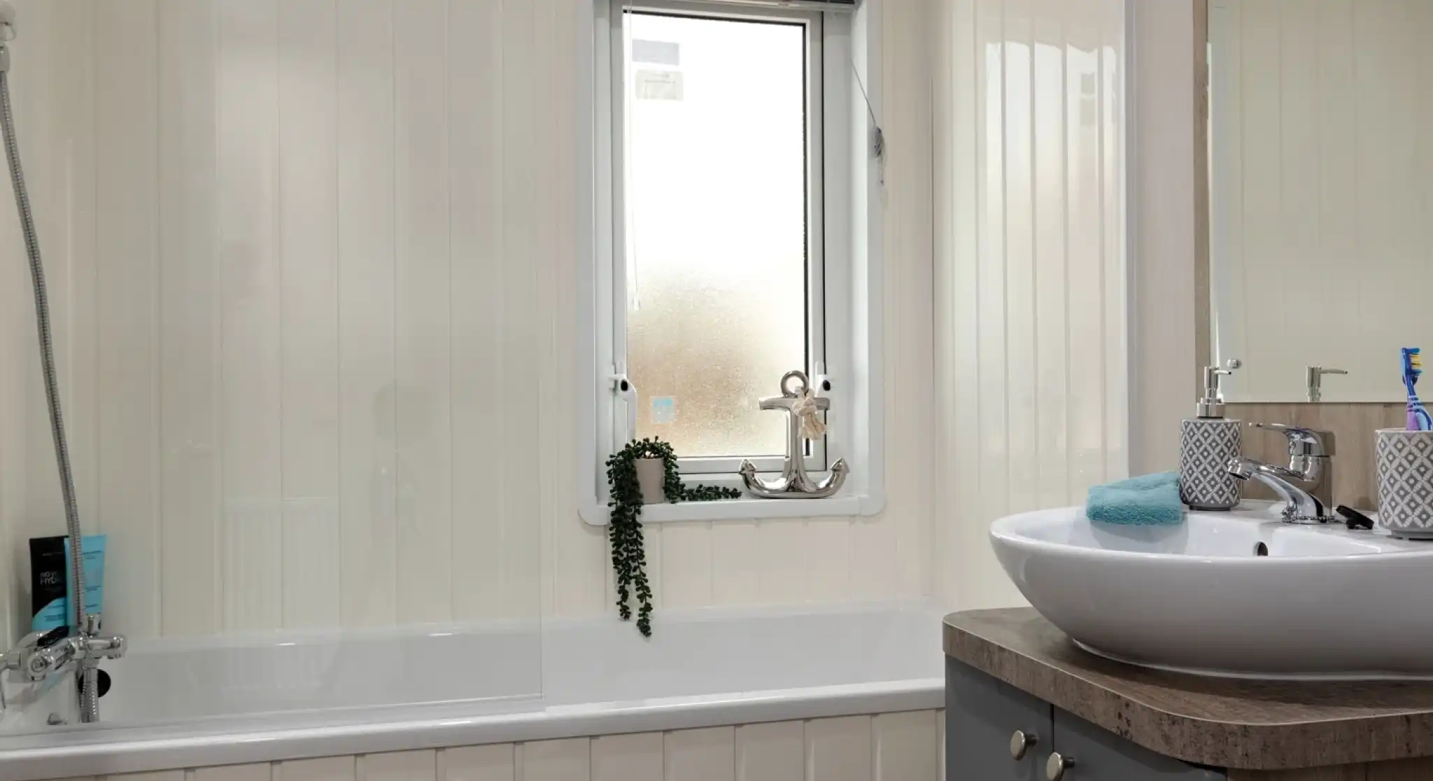 Modern bathroom with a white tub and frosted window. A leafy plant hangs by the window. Sleek sink on a wooden vanity, giving a clean, tranquil feel.
