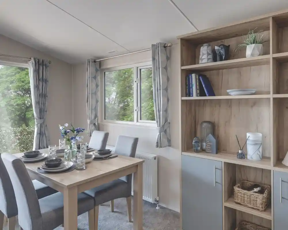 Bright dining area with a wooden table set for four, including grey chairs, plates, glasses, and a flower centerpiece. A wooden shelving unit beside the table displays decorative items, books, and a small plant. The room has light grey curtains and a window with a view of greenery outside.