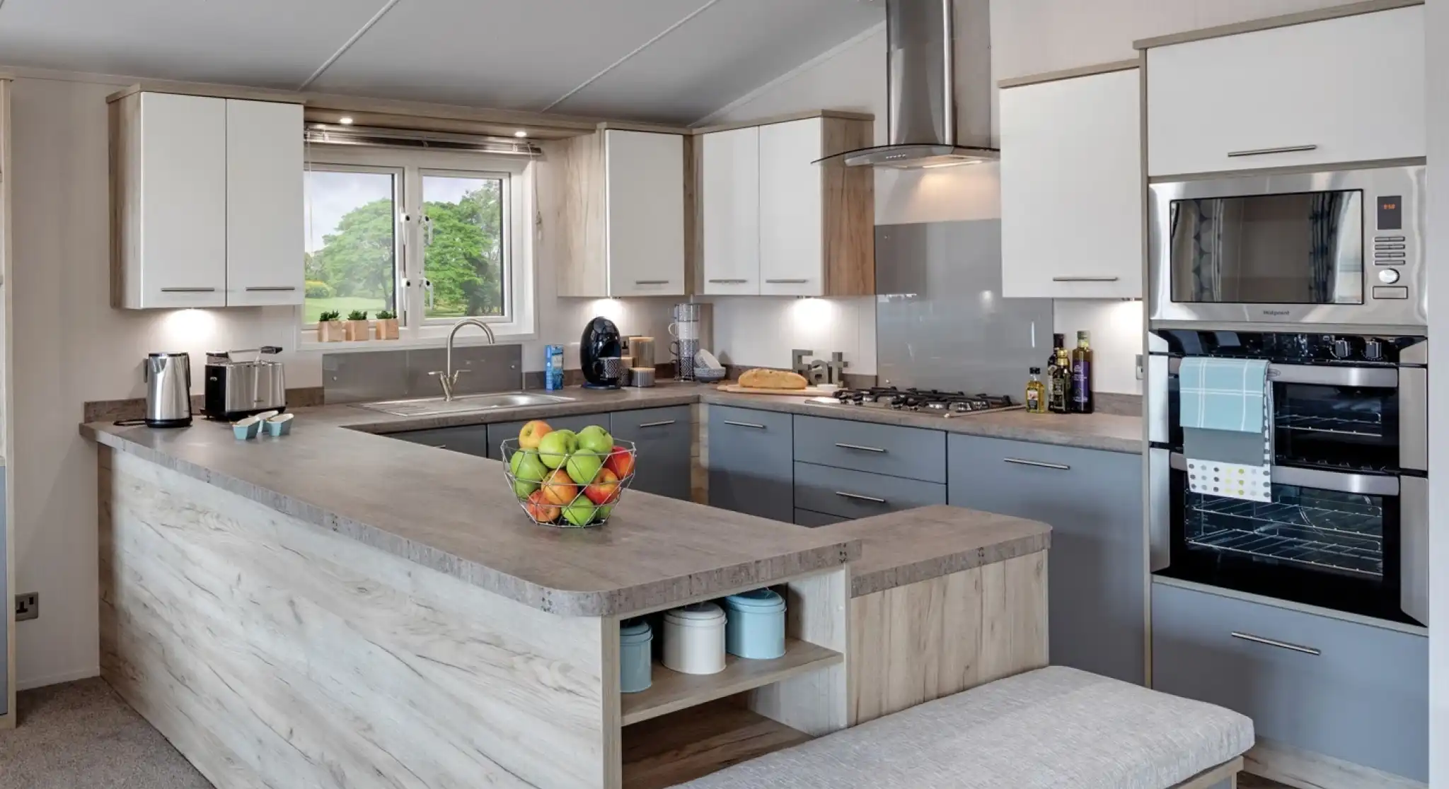 Modern kitchen with a large grey-and-wood island, white and grey cabinetry, stainless steel appliances including a microwave and oven, and a large window offering a view of the outdoors. A bowl of fresh fruit is placed on the countertop, and kitchen essentials like coffee makers and olive oil bottles are visible.