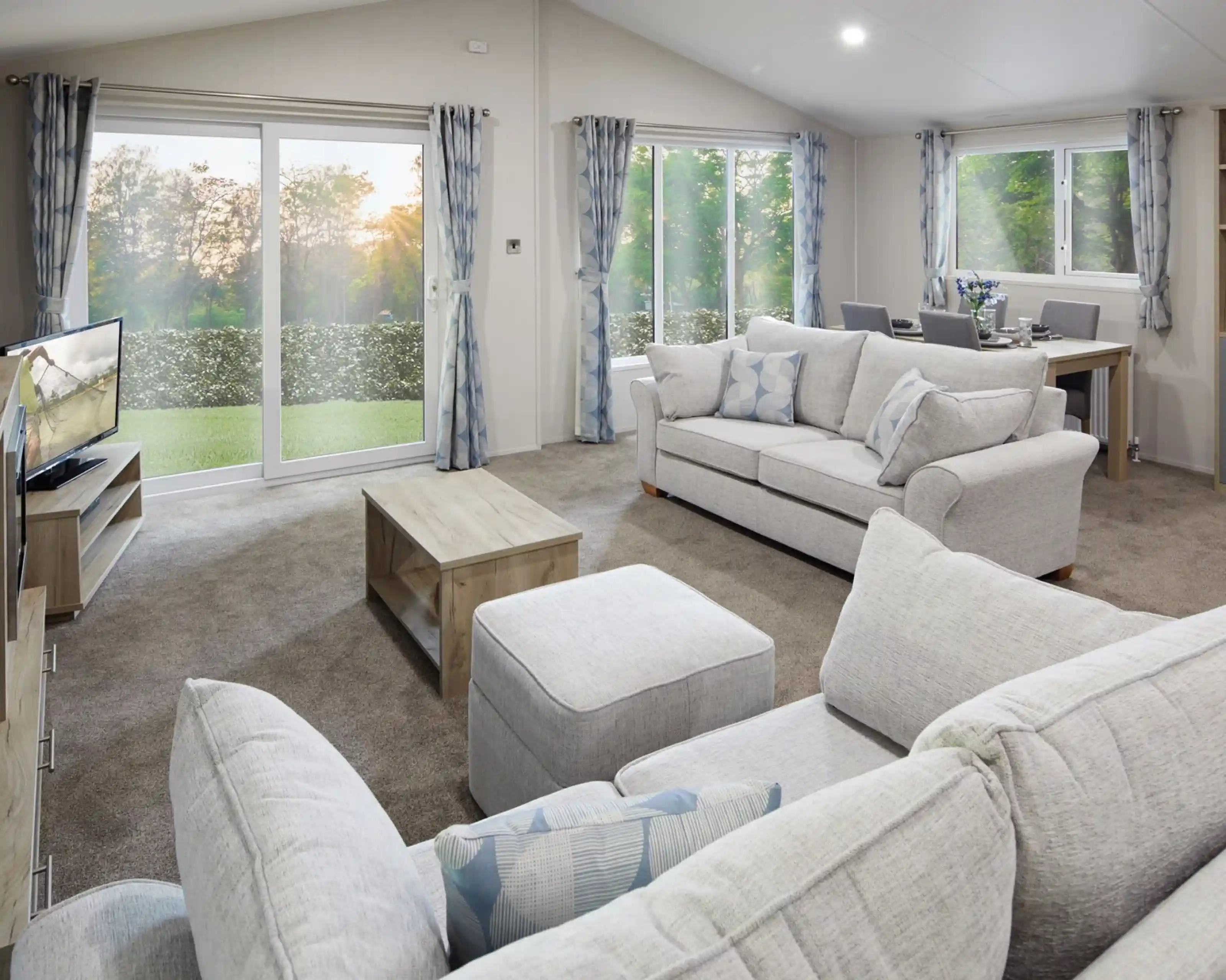Modern living room with light grey sofas, matching curtains, a flat-screen TV, wooden coffee table, and dining area in the background. Large sliding glass doors and windows let in natural light and offer a view of a green outdoor landscape at sunset.
