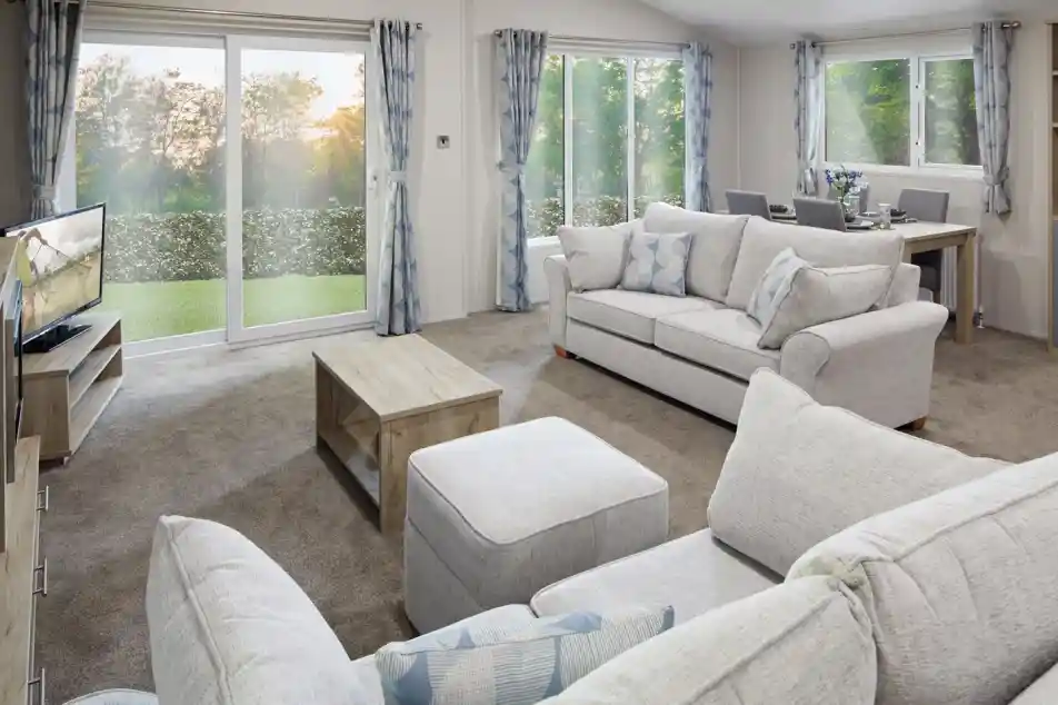 Modern living room with light grey sofas, matching curtains, a flat-screen TV, wooden coffee table, and dining area in the background. Large sliding glass doors and windows let in natural light and offer a view of a green outdoor landscape at sunset.