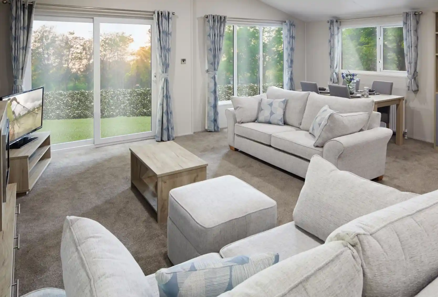 Modern living room with light grey sofas, matching curtains, a flat-screen TV, wooden coffee table, and dining area in the background. Large sliding glass doors and windows let in natural light and offer a view of a green outdoor landscape at sunset.