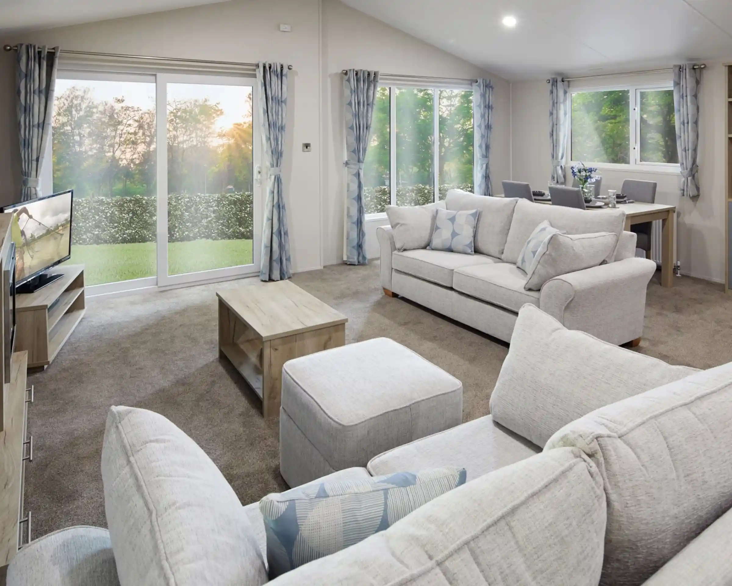 Modern living room with light grey sofas, matching curtains, a flat-screen TV, wooden coffee table, and dining area in the background. Large sliding glass doors and windows let in natural light and offer a view of a green outdoor landscape at sunset.