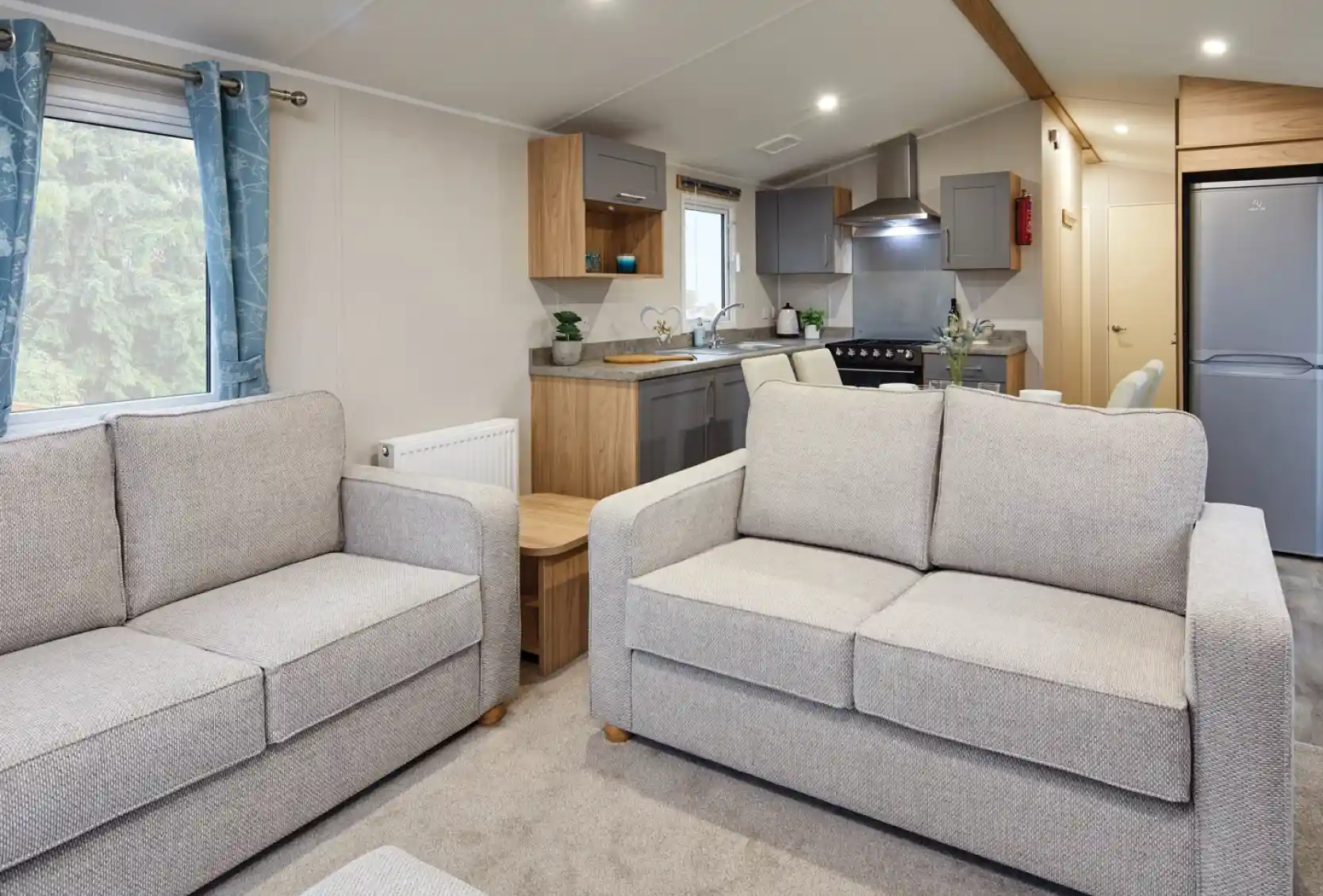 Two grey sofas in a modern living room and kitchen in the background with free-standing fridge freezer, grey cabinetry, stove and sink.