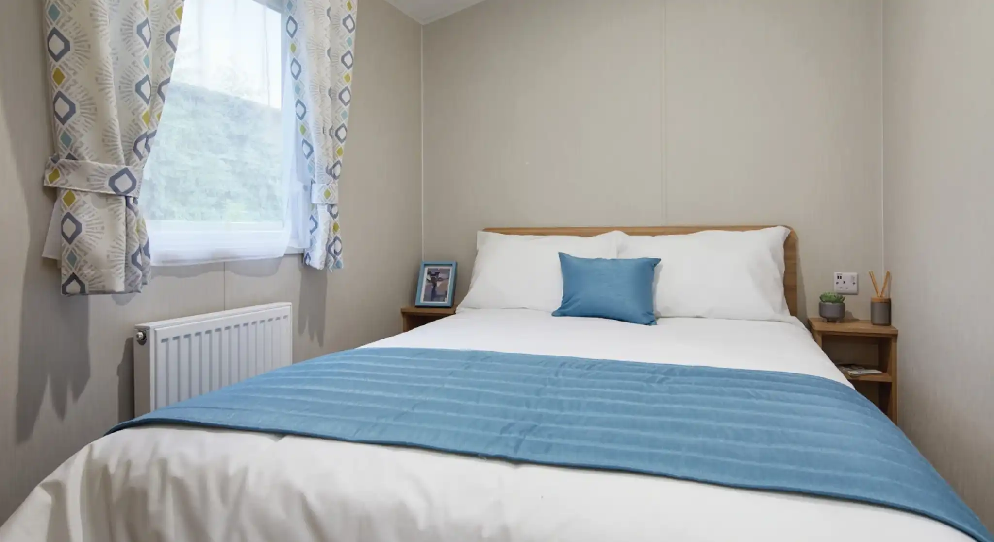 Bright master bedroom in a holiday caravan with white linens, blue accents, light wood furniture, and soft neutral décor for a calm, inviting feel.