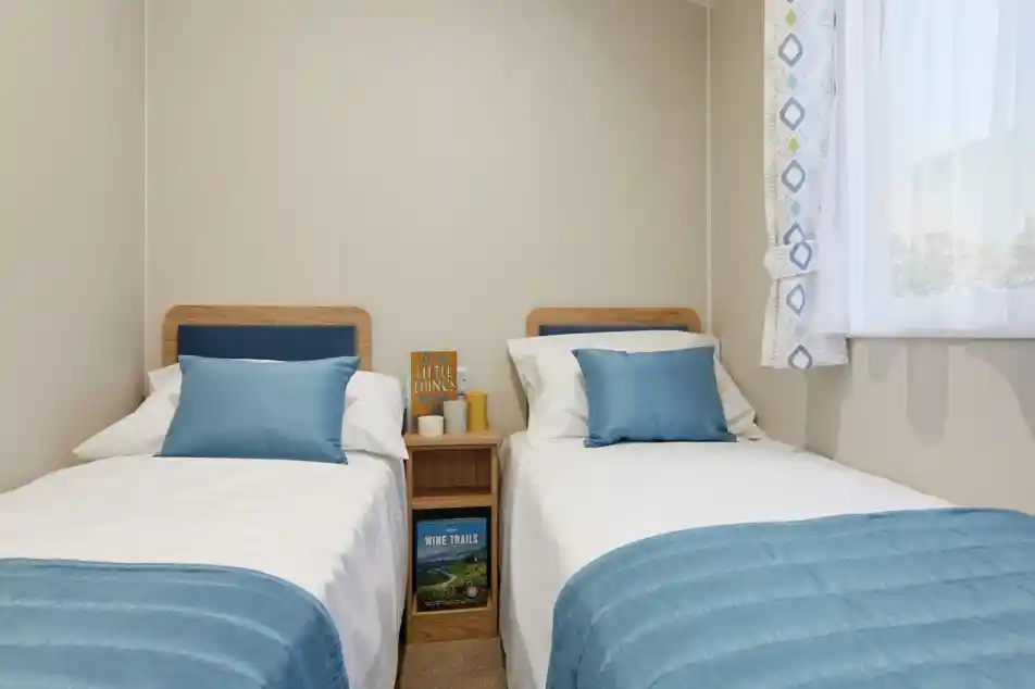 Twin bedroom in a holiday caravan with two single beds, white linens, blue accents, light wood furniture, and a bright, airy feel.