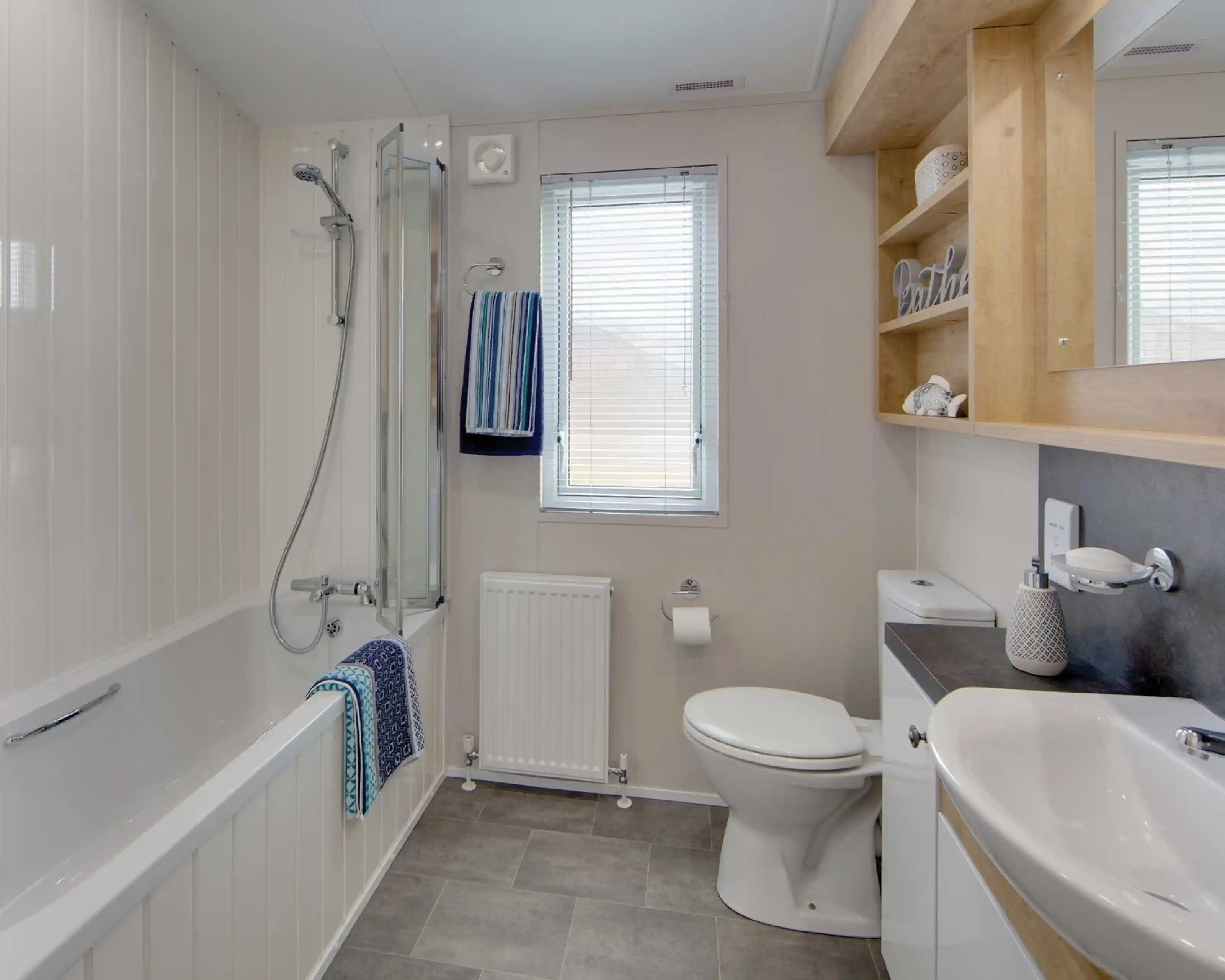 Modern bathroom with gray tile floor, white walls, and a bathtub with glass shower screen. Towels hang near a window above a radiator.