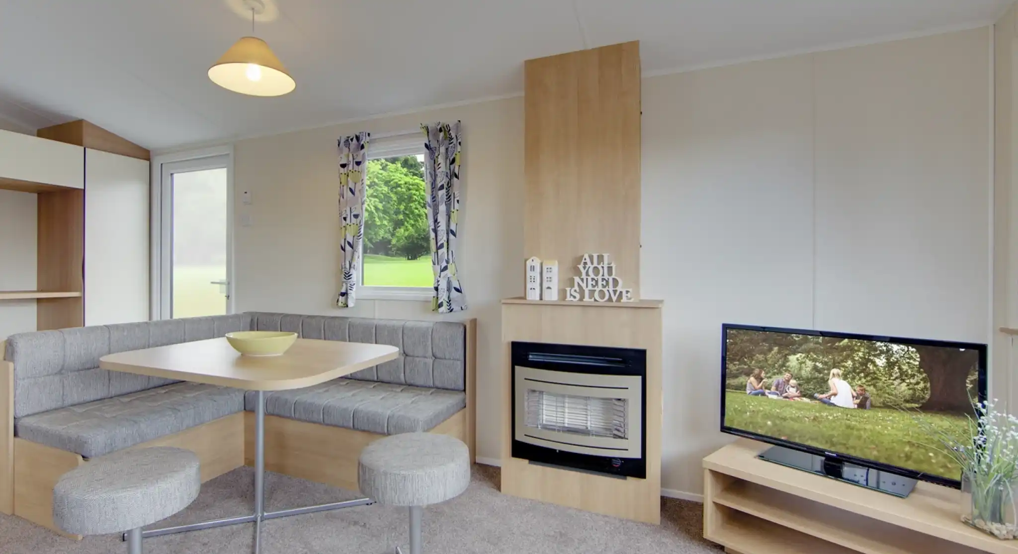 A caravan lounge with a dining banquette, fireplace, and television displaying a family picnic scene.